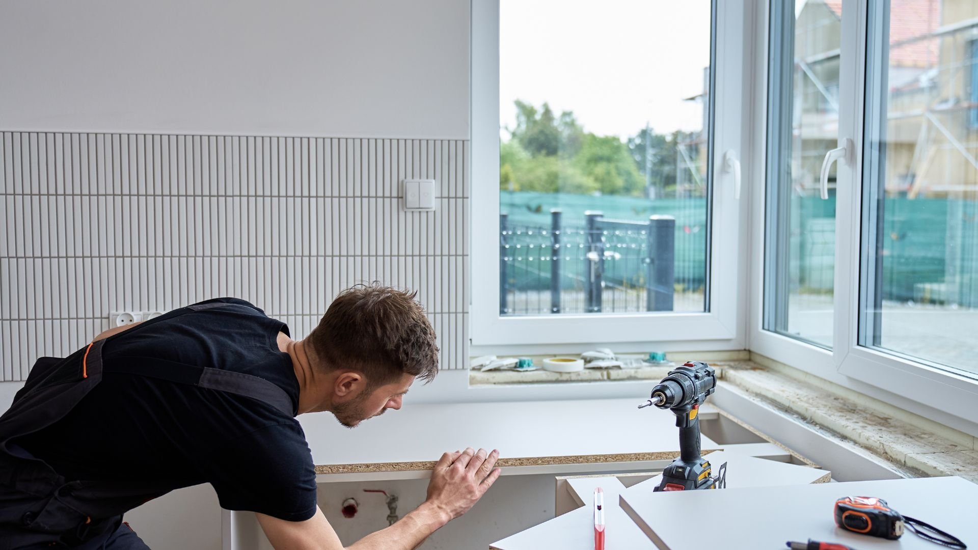 Worker installing kitchen cabinets near window with power drill